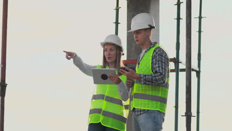 Two-engineers-a-man-and-a-woman-in-white-helmets-with-a-tablet-computer-at-the-construction-site-condemn-the-construction-plan-of-the-building